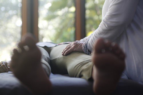 A practitioner's hand resting on the hip of a client lying on a healing table beside a window full of greenery